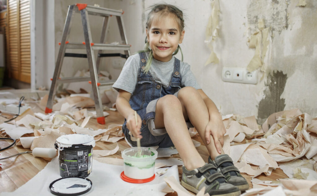 A young girl in dungarees sits on the floor surrounded by torn wallpaper, holding a paintbrush and a container. A stepladder and messy renovation supplies are visible in the background. The girl is smiling at the camera.