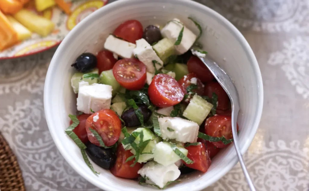 A bowl of Greek salad with cherry tomatoes, cucumbers, black olives, cubed feta cheese, and chopped fresh herbs, with a spoon resting inside the bowl.