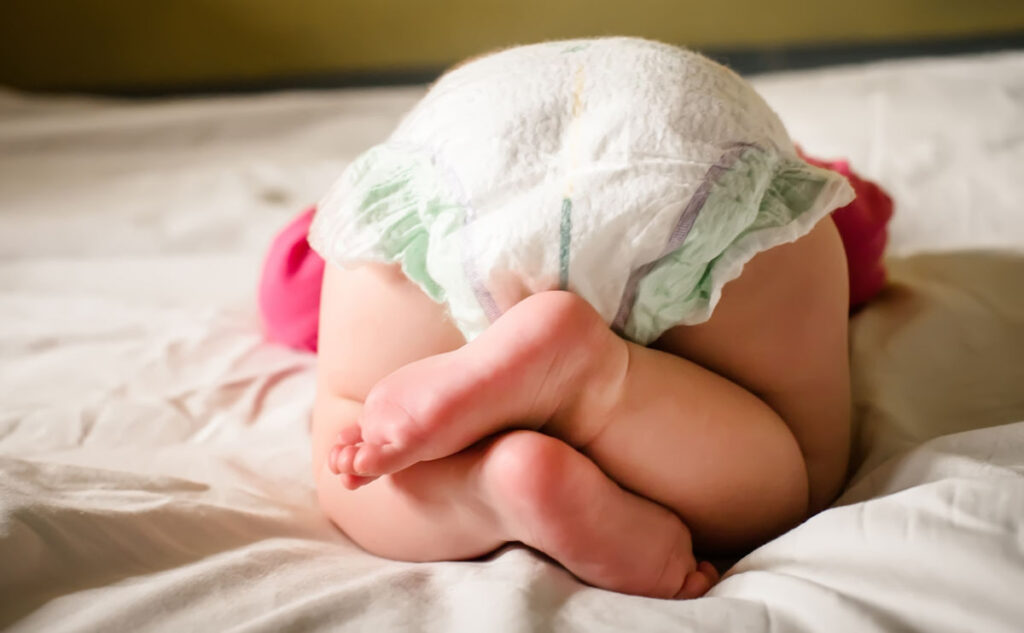 A baby in a diaper is lying on a bed with legs crossed and facing away from the camera, showing their back and feet.