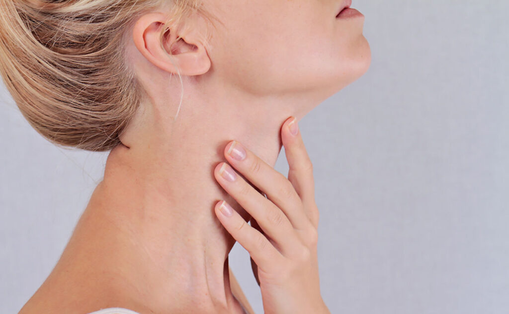 A woman tilts her head back and touches her neck gently with her hand, showing a close-up of her jawline and throat against a plain, light background.