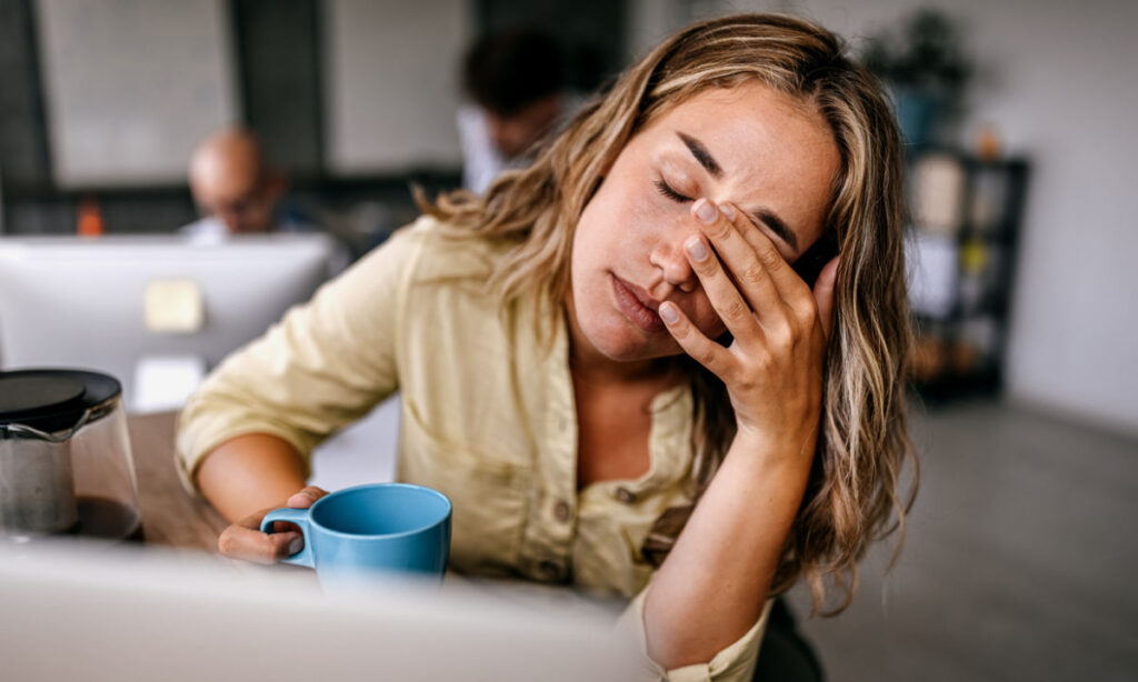 A woman with long hair sits at a desk holding a blue mug, looking tired with her eyes closed and one hand covering her face. There are computers and other people in the blurred background.