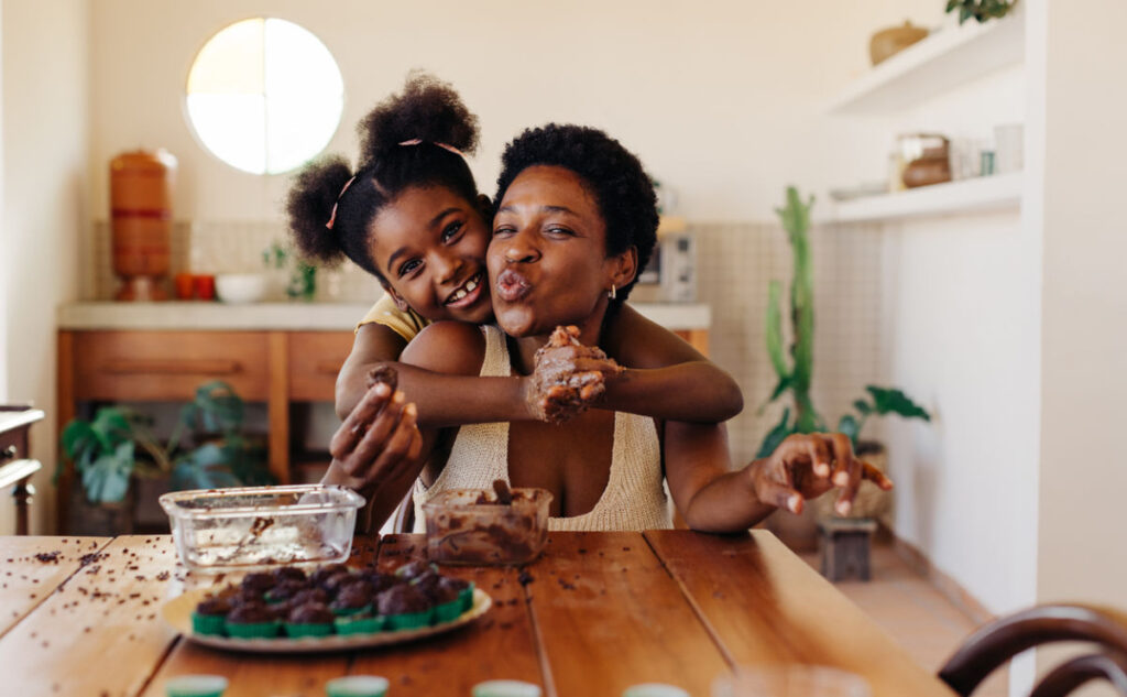 A young girl hugs a woman from behind as they smile and make silly faces while baking in a kitchen. The table is covered with chocolate treats and baking ingredients.