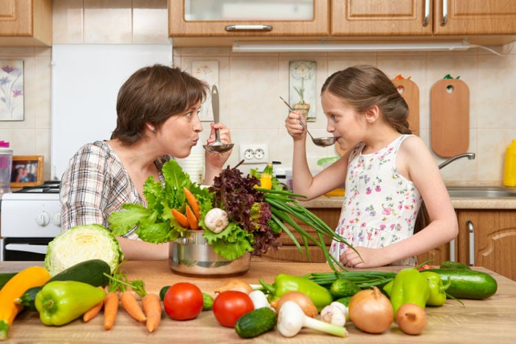 A woman and a young girl playfully point forks at each other over a bowl of salad in a kitchen, surrounded by fresh vegetables like tomatoes, peppers, cucumbers, and lettuce on the worktop.