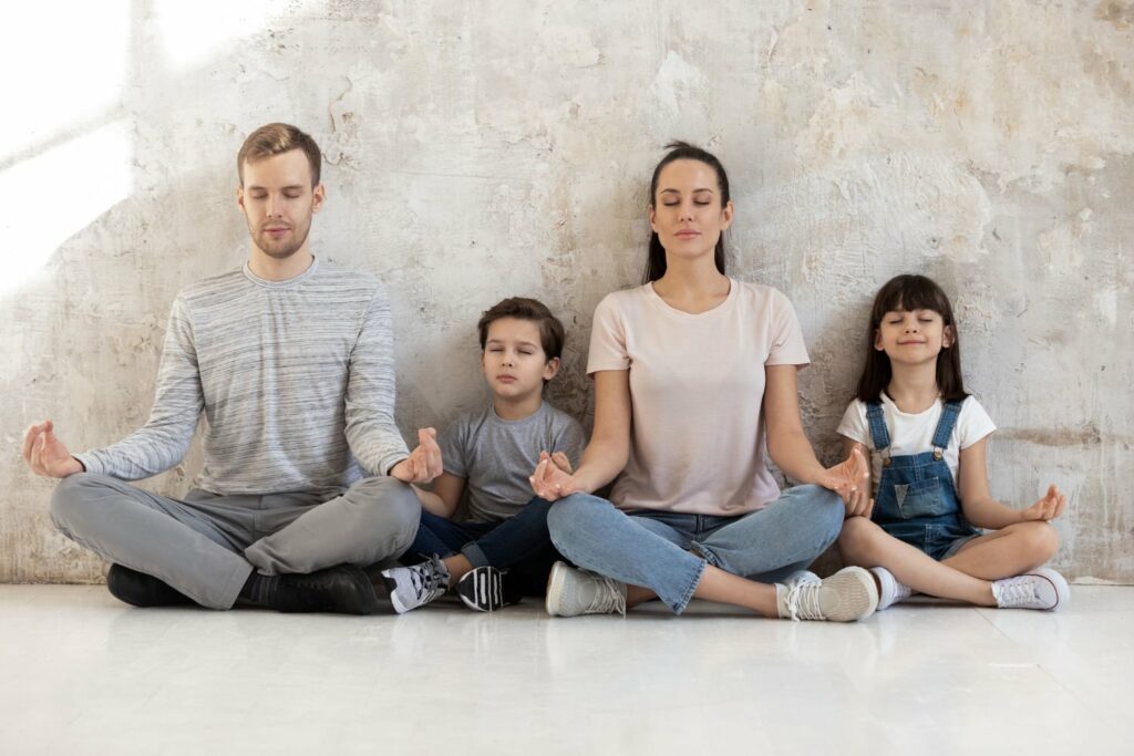 A family of four—two adults and two children—sit cross-legged on the floor against a textured wall, eyes closed and hands in meditation poses, appearing calm and relaxed.