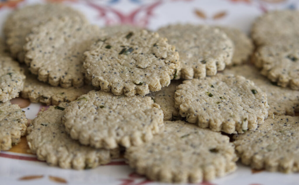 A close-up of round, scalloped-edge crackers with speckled herbs, arranged in a pile on a decorative plate. The crackers appear homemade with a rustic texture.