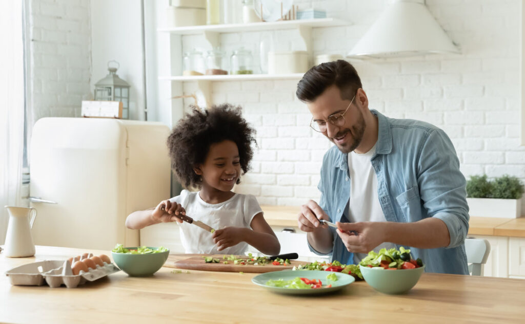 A man and a young girl are smiling while preparing a salad together in a bright kitchen. The countertop is filled with fresh vegetables and eggs. They appear to be enjoying the cooking activity.