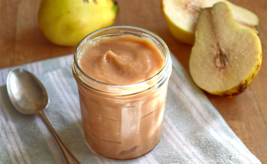 A jar of creamy quince purée on a striped cloth next to a spoon. Fresh quinces, with one halved to reveal the seeds, are in the background on a wooden surface.