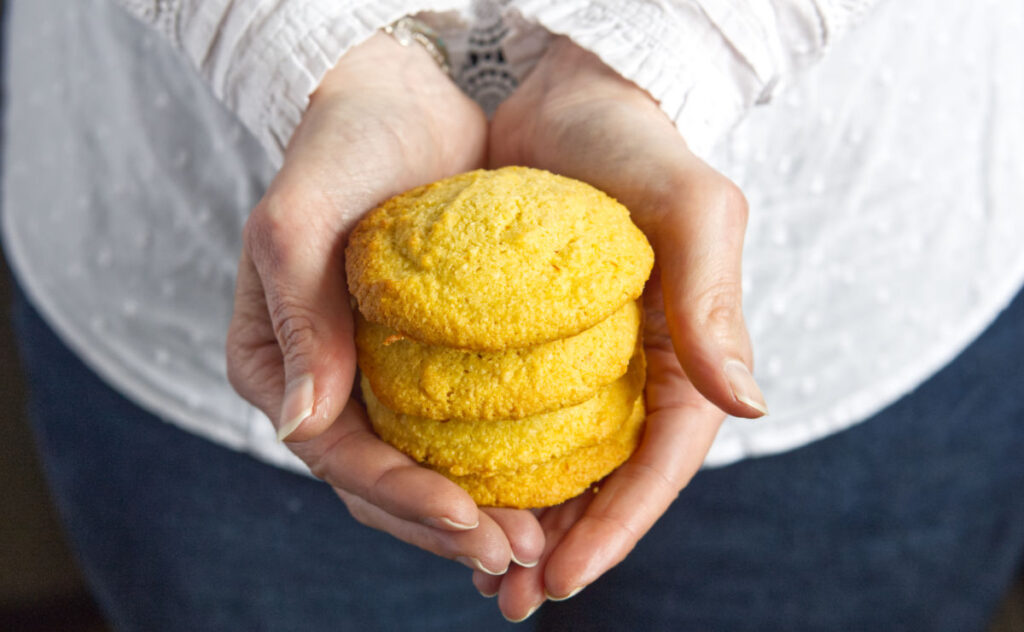 Hands holding a stack of four yellow golden-brown cookies. The person is wearing a white long-sleeve shirt and blue jeans. The cookies appear freshly baked, with a soft and crumbly texture.