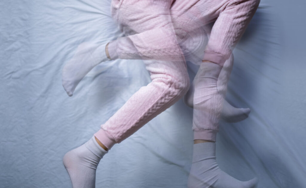 A blurred image showing a child in pink pajama pants and white socks lying on a bed. The double exposure effect creates an impression of movement, with legs slightly overlapping in different positions.