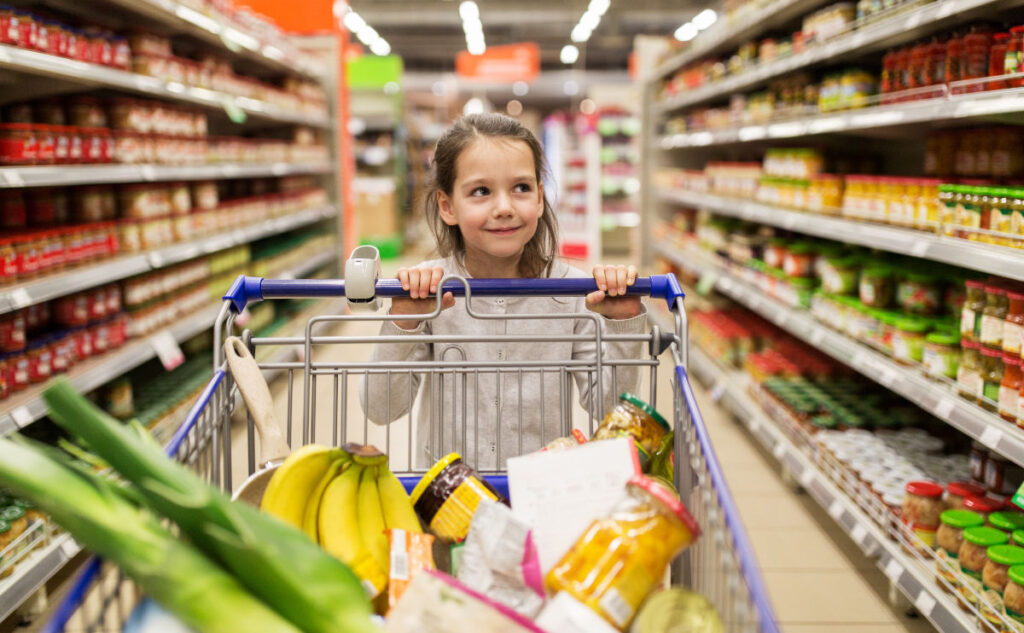 A young girl pushes a supermarket cart filled with various items, including bananas and jars. She is smiling and looking to the side, surrounded by aisles of stocked shelves in a supermarket.