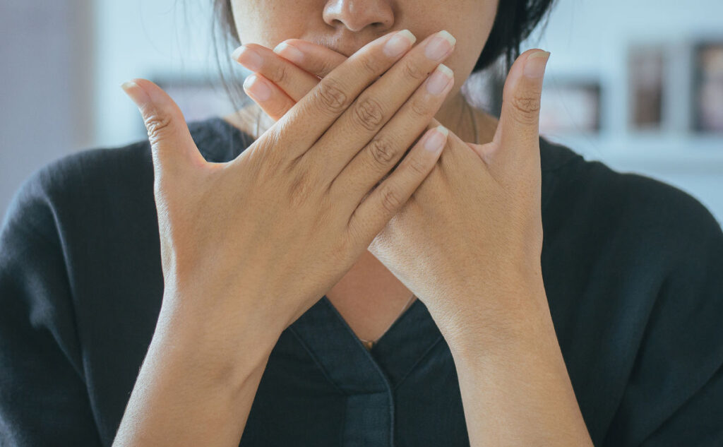 A person in a dark shirt covers their mouth with both hands, displaying neatly trimmed nails. The background is blurred, focusing attention on the gesture.