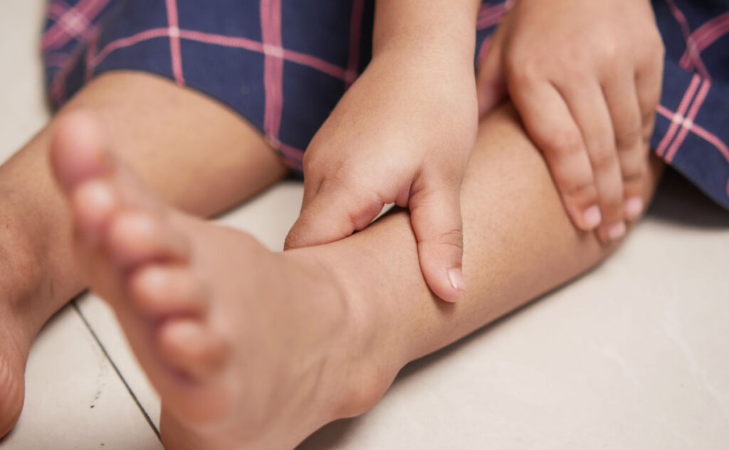 A child sitting on the floor with a hand pressing against the lower leg, indicating pain or discomfort. The child is wearing a garment with a plaid pattern.