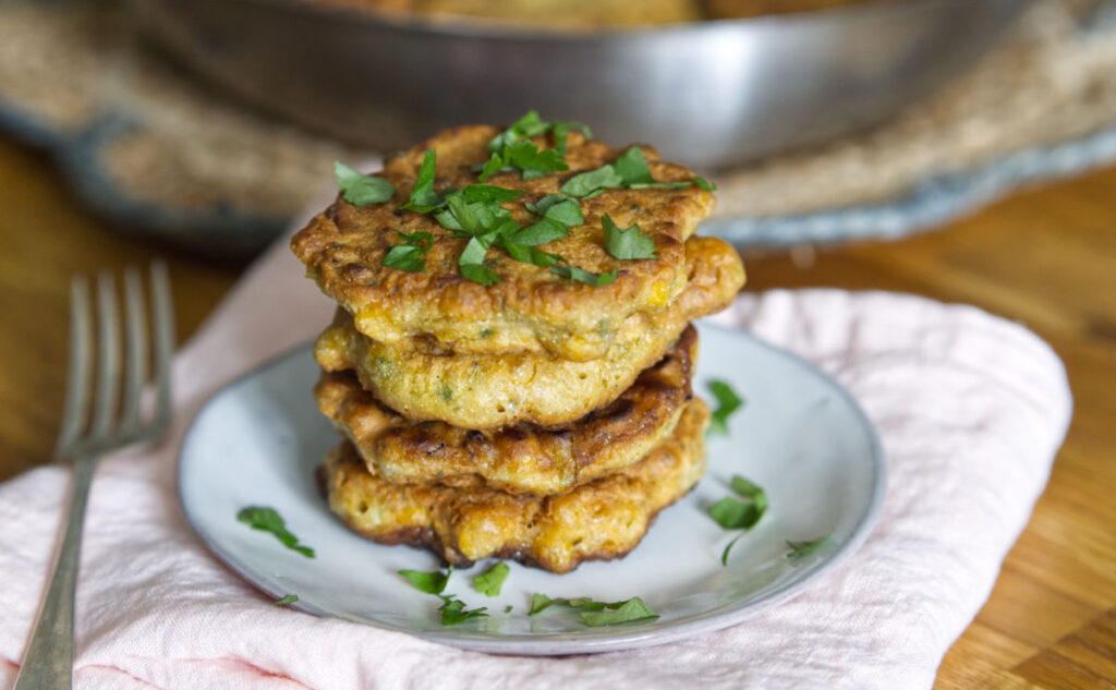 A stack of three Sweetcorn and Coriander Fritters garnished with chopped parsley on a white plate. The plate rests on a soft white cloth, with a fork beside it. The background shows the pan they were cooked in and a wooden table surface.