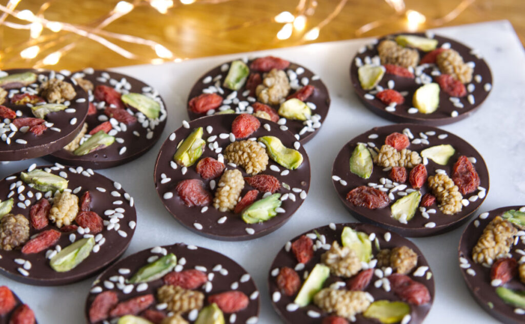 Round pieces of dark chocolate topped with pistachios, goji berries, mulberries, and sesame seeds are arranged on a white surface. Delicate string lights are softly glowing in the background.