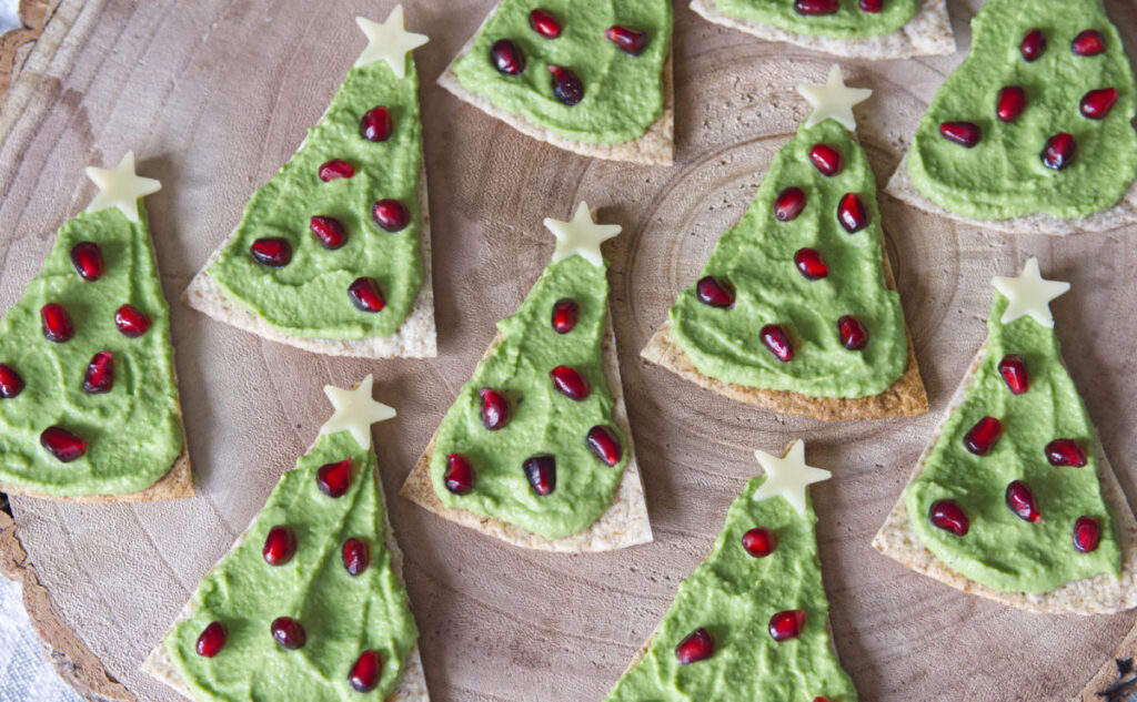 Festive tortilla chips topped with green guacamole, decorated with red pomegranate seeds and star-shaped cheese pieces, arranged on a wooden surface to resemble Christmas trees.