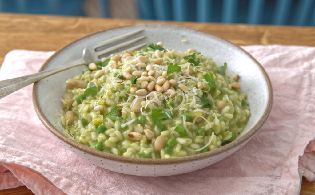 A bowl of creamy risotto topped with pine nuts and fresh parsley. The dish rests on a pink cloth, with a fork placed on the side, and a blue chair visible in the blurred background.