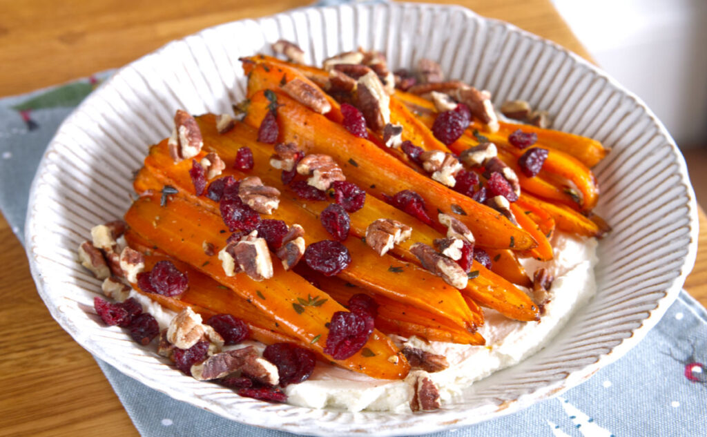 A bowl of roasted carrots on a bed of creamy cheese, topped with chopped pecans and dried cranberries. The dish is set on a decorative cloth on a wooden table.