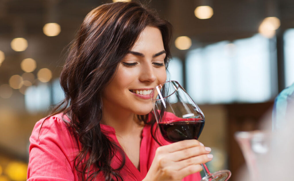 Woman in a red blouse smiling while holding a glass of red wine, appearing to enjoy its aroma. But is she about to sneeze? She is indoors with blurred lights in the background.
