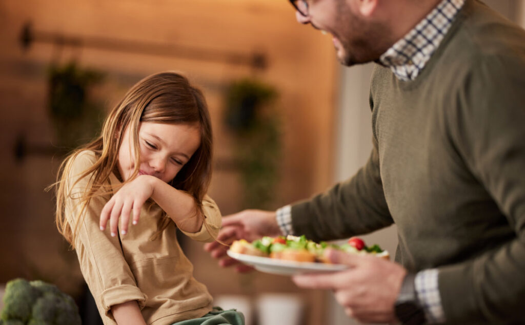 A little girl recoils as she covers her mouth with her arm to refuse the food she is offered by her father. He, beside her, wearing glasses and a green sweater, holds a plate of salad. They appear to be in a cozy kitchen setting.