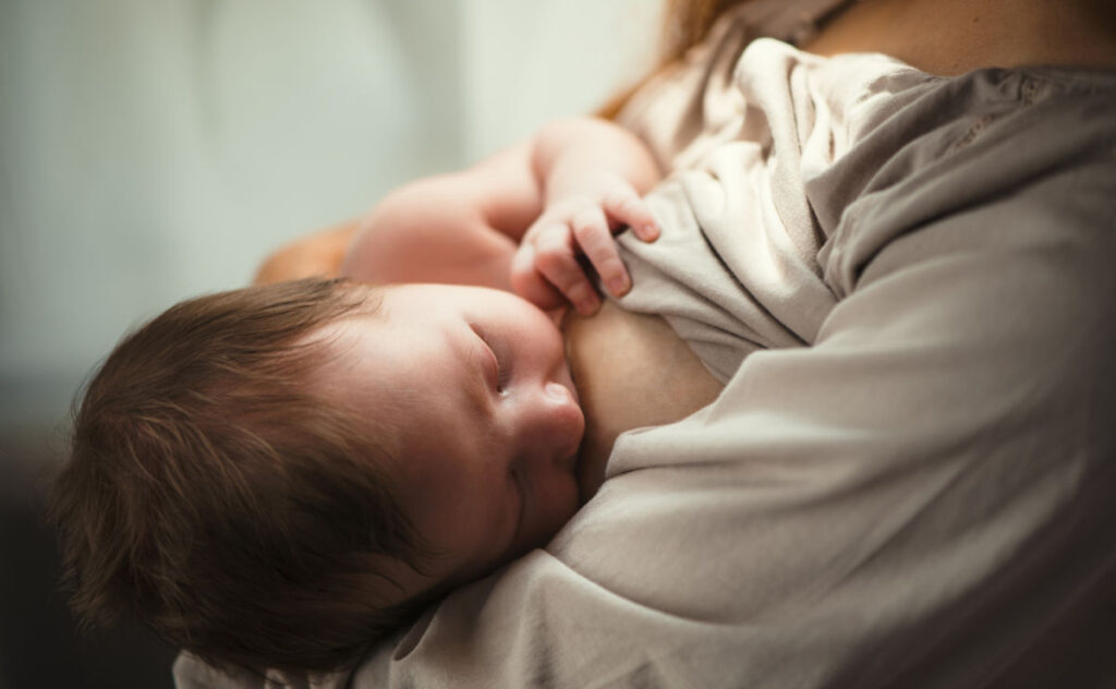 A baby with dark hair is breastfeeding, cradled in the arms of an adult wearing a light-coloured top. The baby’s hand rests gently on the adult’s chest.