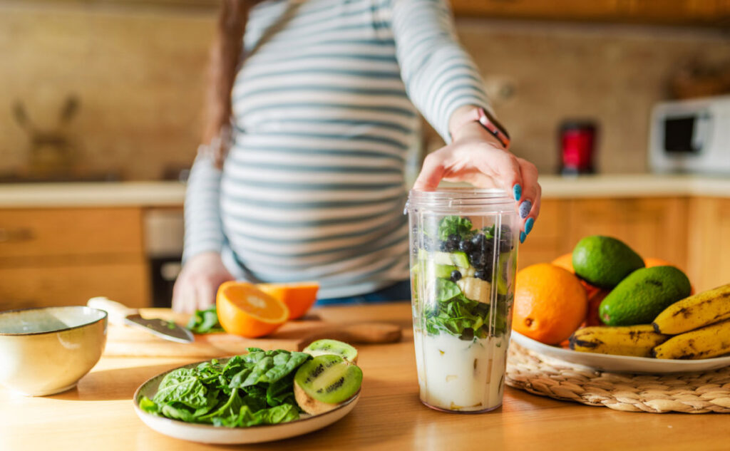 A pregnant woman in a striped shirt stands in a kitchen, preparing a smoothie with spinach, banana, kiwi, and berries. Fresh fruit and spinach are arranged on the worktop.