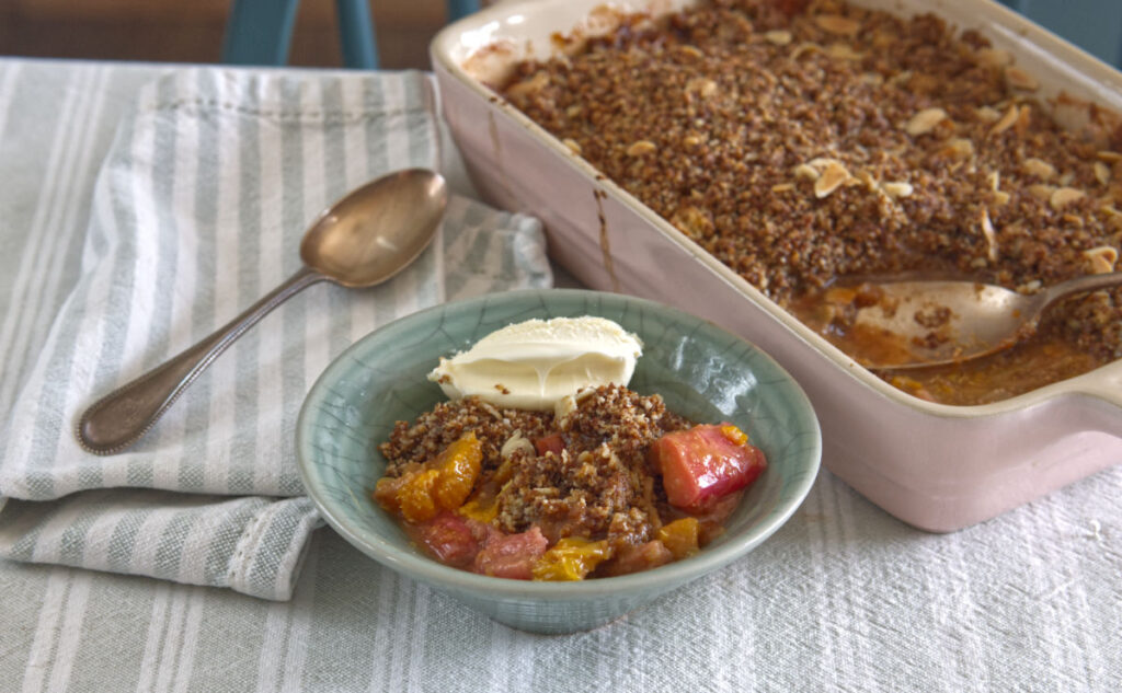 A bowl of fruit crumble topped with a dollop of cream sits on a striped cloth next to a spoon. A larger dish of crumble with fruit and a crumbly topping is in the background. The tablecloth is light gray with a simple pattern.