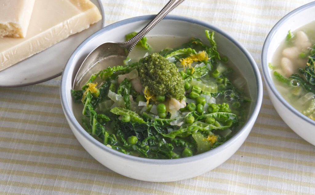 A bowl of vegetable soup with leafy greens, peas, white beans, and a dollop of green pesto on top, served with a spoon. A plate with a wedge of cheese is visible in the background.