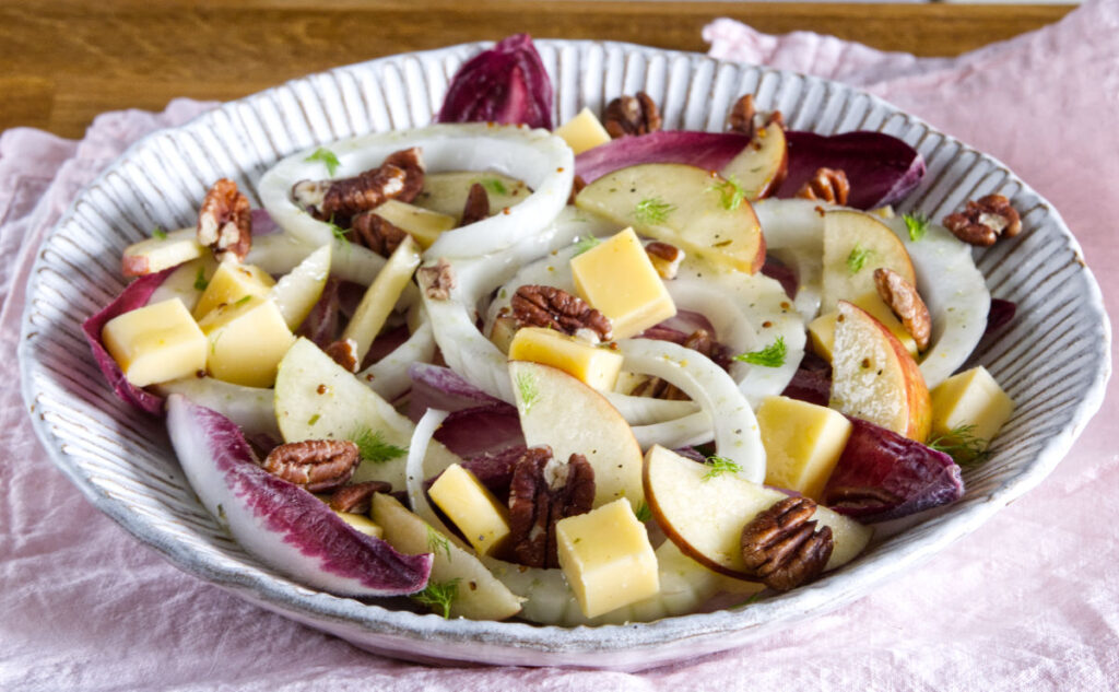 A salad in a white bowl with apple slices, fennel, cubed cheese, radicchio leaves, and pecans, served on a pink cloth.