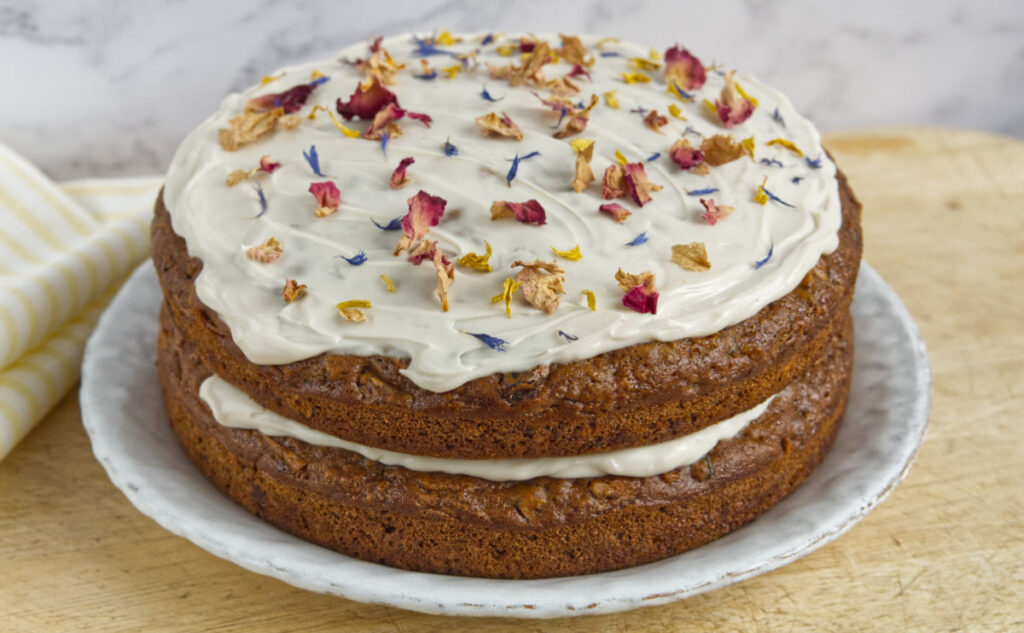 A two-layer cake with creamy white frosting between the layers and on top, decorated with colorful dried flower petals, sits on a white plate on a wooden surface.
