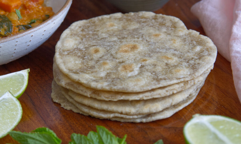 A stack of four flatbreads sits on a wooden surface, surrounded by lime wedges, mint leaves, and a bowl of curry. The flatbreads appear soft and lightly browned.