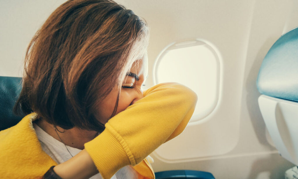 A woman sitting on an airplane sneezes or coughs into their bent elbow, wearing a yellow sweater, with a window and seat visible in the background.