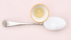 A small bowl of cooking oil and a metal spoon filled with baking powder are placed on a light pink surface.