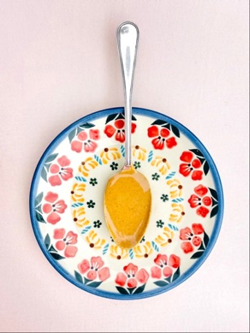 A silver spoon with peanut butter rests on a colourful floral-patterned plate with red, yellow, and green designs, set on a light pink background.