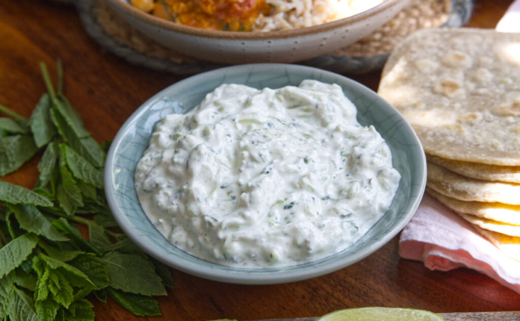 A bowl of raita, a creamy yogurt-based Indian condiment with chopped herbs, sits on a wooden table next to fresh mint leaves, flatbreads, a slice of lime, and a plate of rice and curry in the background.