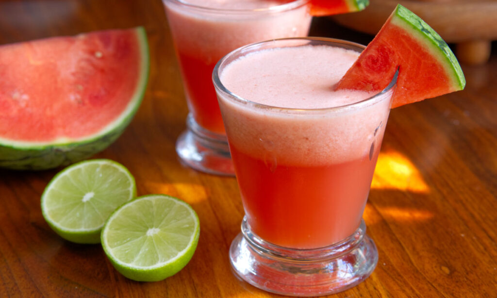 A glass of fresh watermelon juice with a frothy top, garnished with a watermelon slice. Next to the glass are halved limes and a wedge of watermelon on a wooden surface. Another glass of juice is in the background.