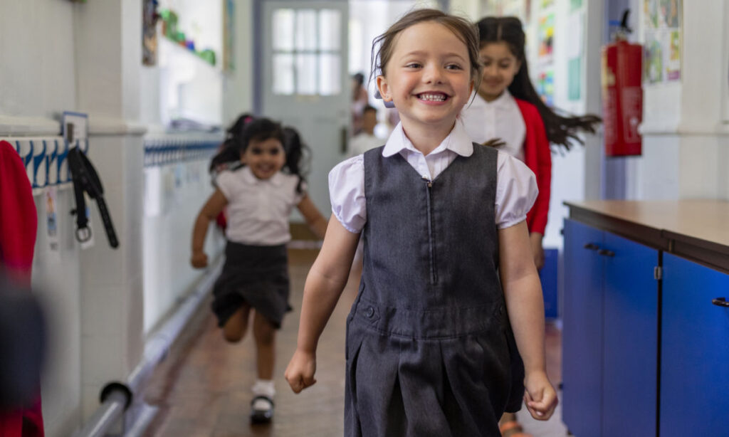 Three young girls in school uniforms are smiling and running down a brightly lit school hallway, with classroom decorations and coats visible in the background.