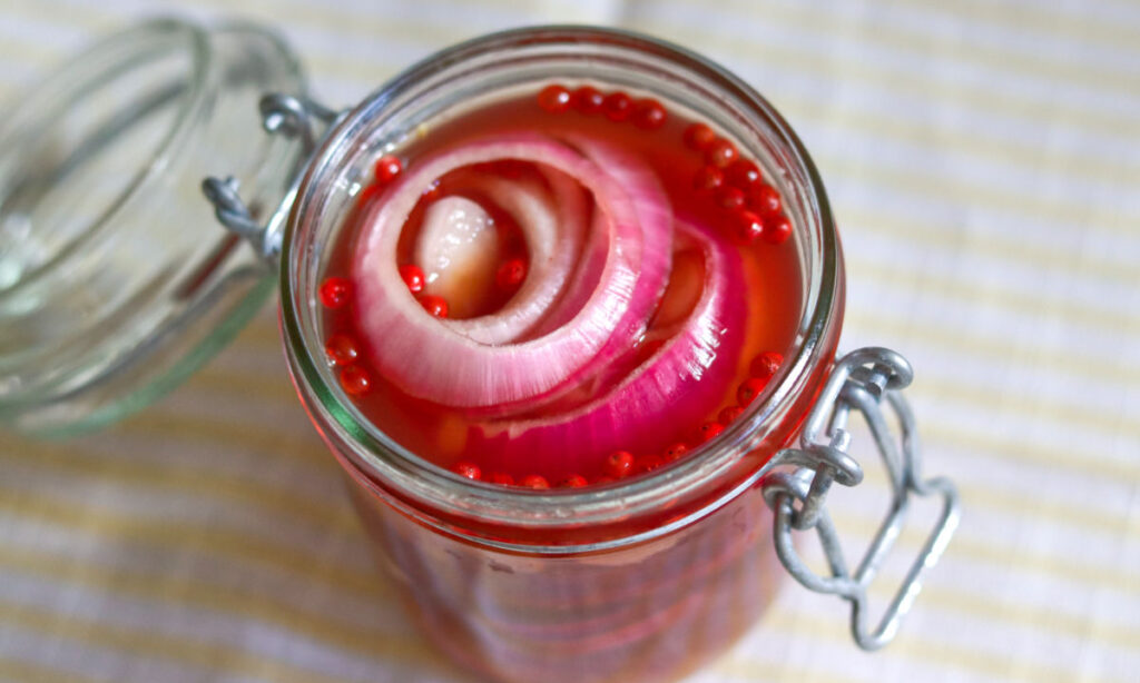 A glass jar filled with pickled red onion slices and red peppercorns in brine, sitting on a yellow and white checkered surface with the lid open.