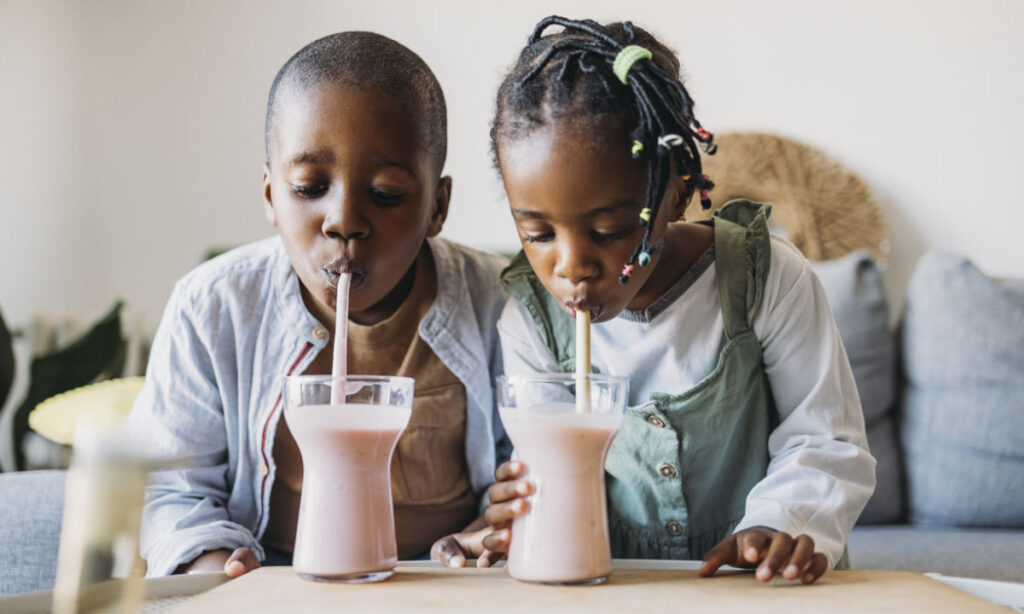 Two children sitting together on a couch, drinking pink smoothies with straws from clear glasses. The boy wears a light shirt and the girl wears a green dress over a white top.