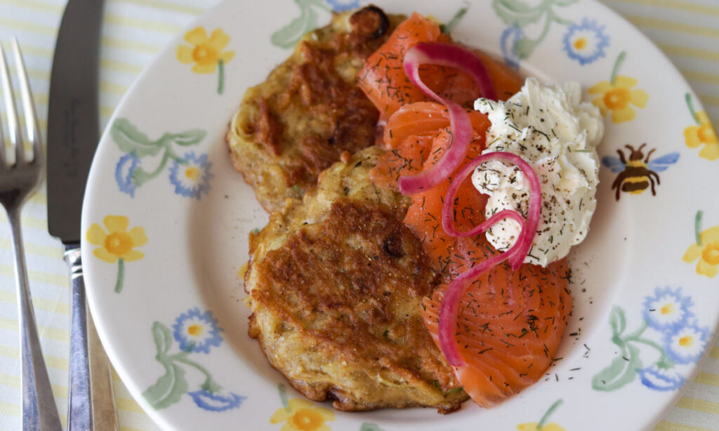 A plate with two golden potato pancakes, slices of smoked salmon, a dollop of cream cheese, pickled red onions, and a sprinkle of dill, on a floral-patterned plate set on a striped tablecloth.