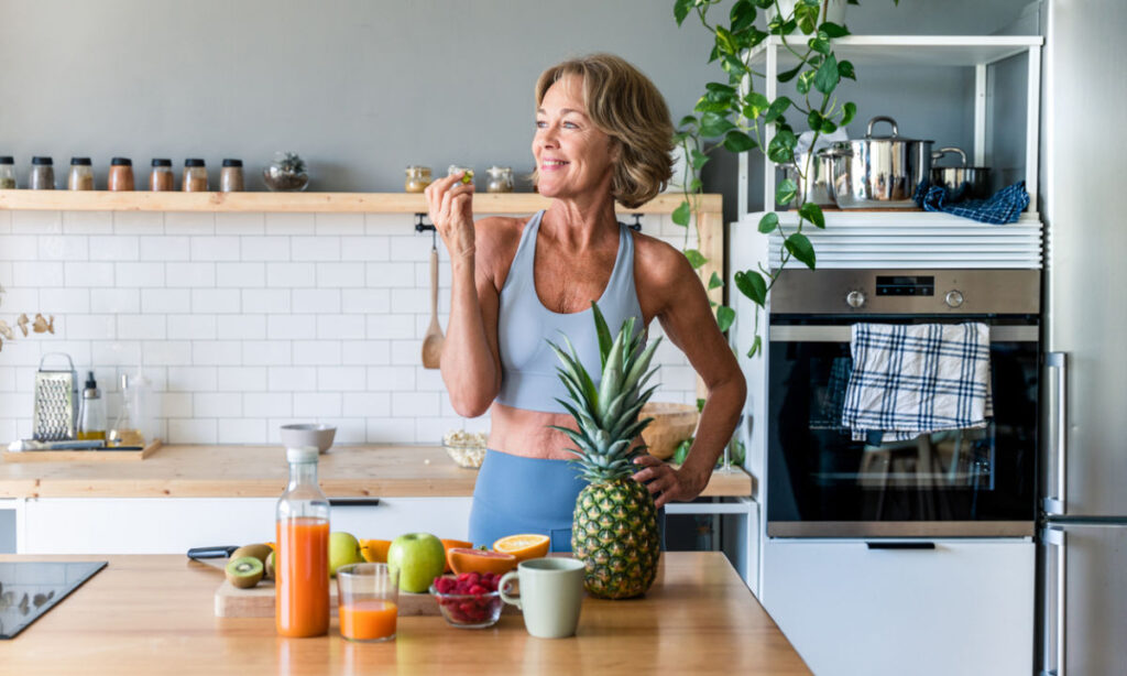 A smiling woman in fitness wear stands in a modern kitchen, eating an apple. A variety of fresh fruits, juice, and a pineapple are on the counter in front of her. The kitchen has open shelves and plants.
