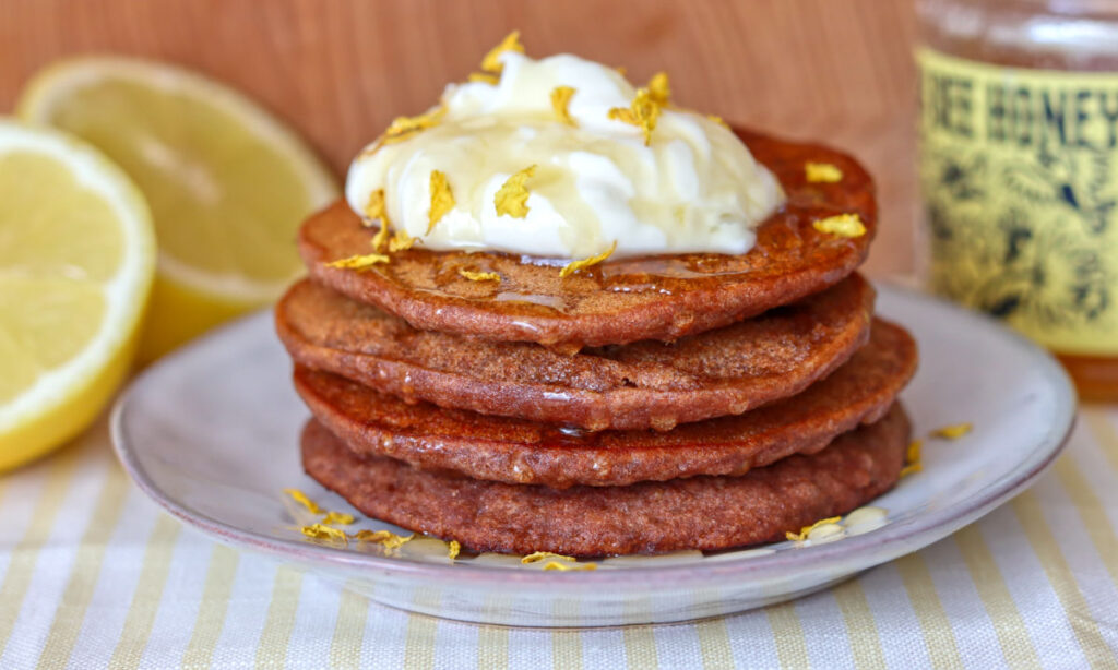 A stack of golden pancakes topped with whipped cream, honey, and yellow flower petals sits on a plate. Lemon halves and a jar of honey are in the background. The scene has a cozy, rustic feel.