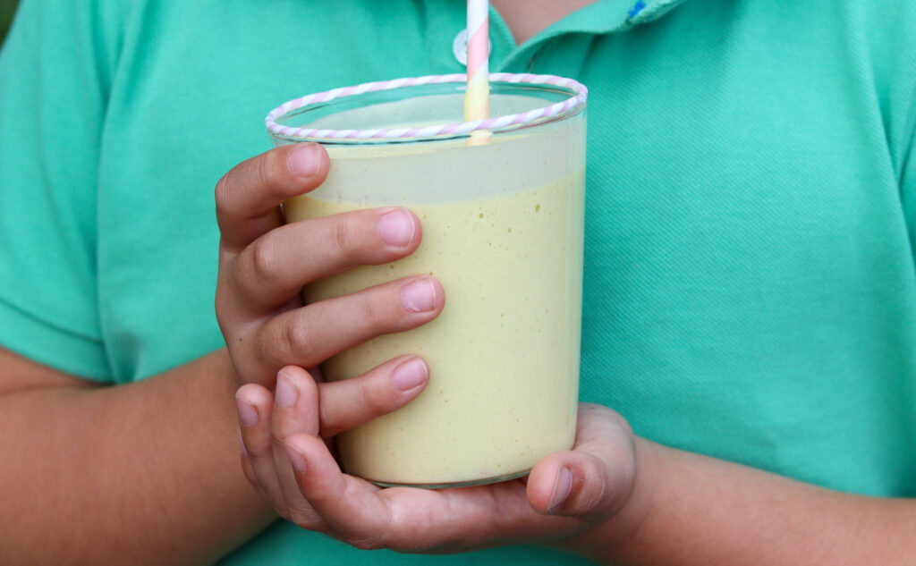 A boy wearing a green shirt holds a glass of Mango Lassi with both hands, drinking from a white and pink striped straw.
