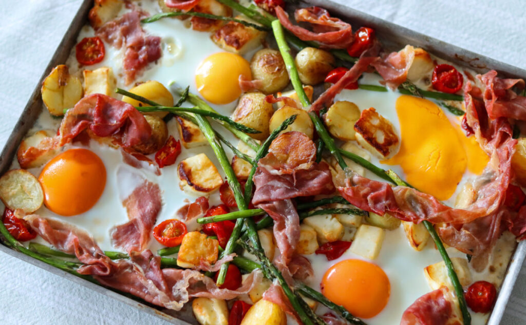 Sheet pan breakfast with baked eggs, crispy prosciutto, roasted potatoes, asparagus, cherry tomatoes, and golden halloumi cheese on a white surface.