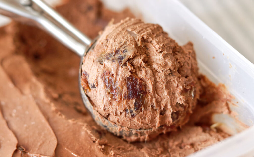 A close-up of a metal ice cream scoop holding a round portion of chocolate ice cream with visible chunks, placed over a container filled with more chocolate ice cream.