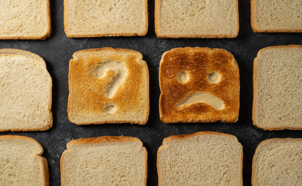 Several slices of bread are laid out on a dark surface. One slice has a question mark shape pressed into it, while another slice is toasted with a sad face, featuring two dots for eyes and a frown.