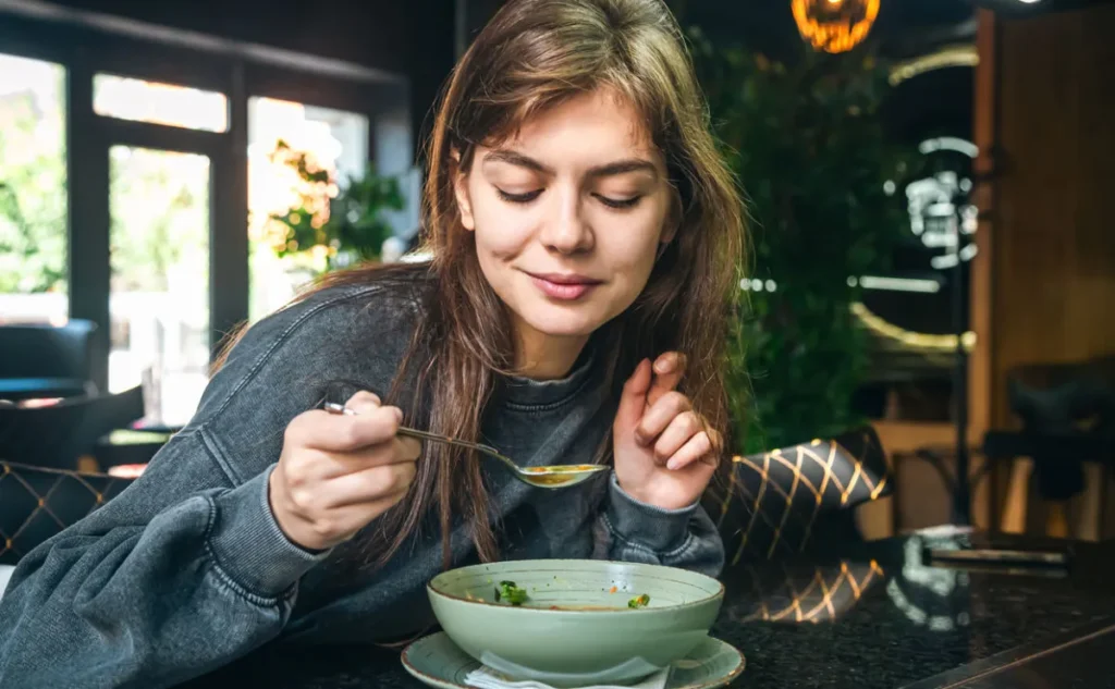 A young woman with long brown hair sits at a table in a restaurant, smiling gently as she holds a spoon and leans over a bowl of soup, full of green vegetables.