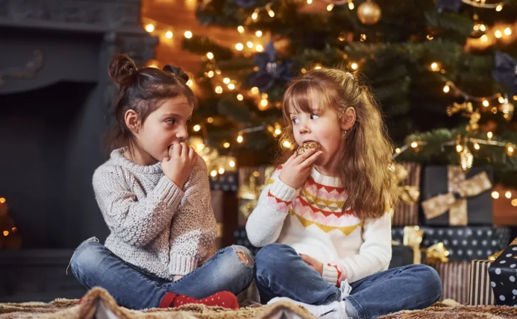 Two young girls sit on the floor in front of a decorated Christmas tree, eating biscuits. They wear cosy jumpers and jeans, and are surrounded by wrapped presents, creating a festive holiday atmosphere.
