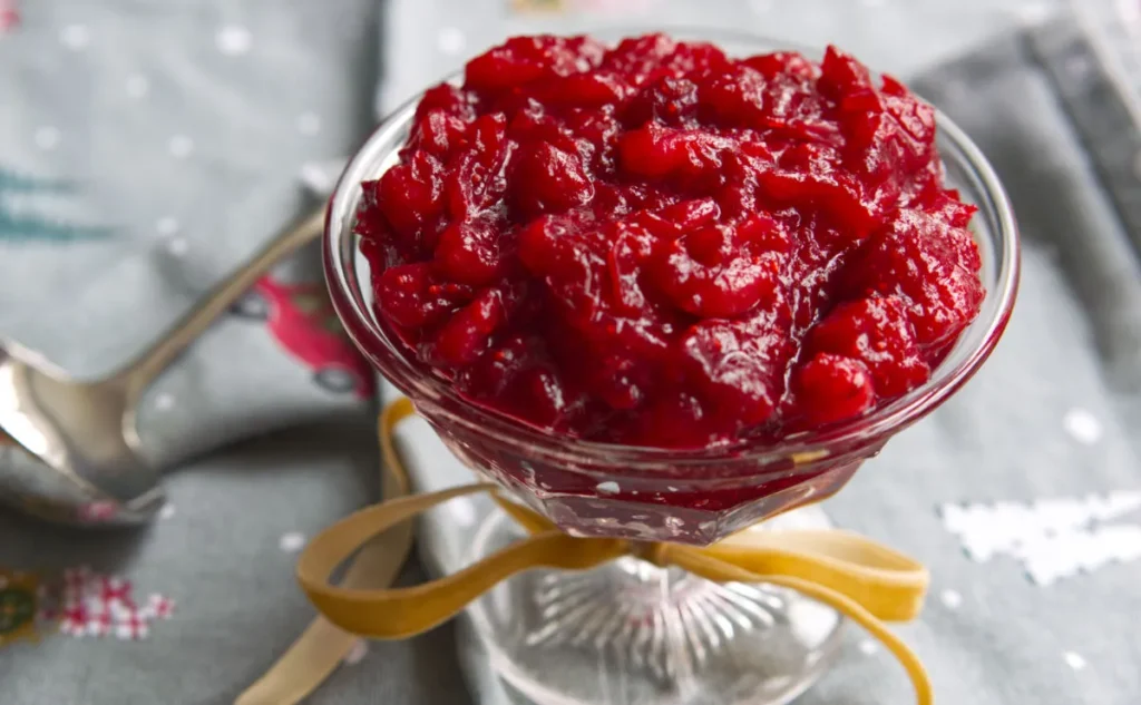 A glass dish filled with chunky, bright red cranberry sauce which sits on a table with a festive grey cloth in the background and a ribbon tied around the dish’s stem. A spoon rests nearby.